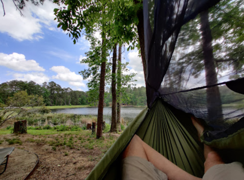 A person relaxes in a hammock with a view of a serene lake and trees under a partly cloudy sky.