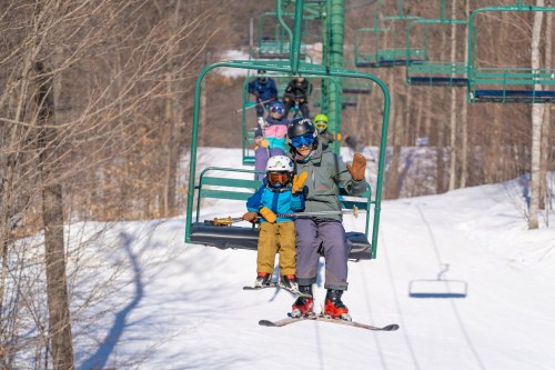 A child and adult ride a ski lift, waving, surrounded by snowy trees and other skiers in the background.