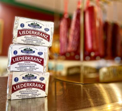 Three packages of Liederkranz cheese stacked on a counter, with hanging sausages blurred in the background.