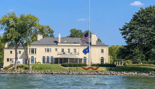 A large, elegant house by the water, featuring an American flag at half-mast and lush green landscaping.