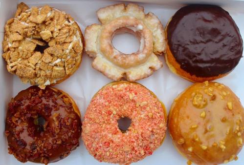 A box of six assorted donuts: glazed, chocolate, cereal-topped, maple bacon, strawberry, and nut-covered varieties.