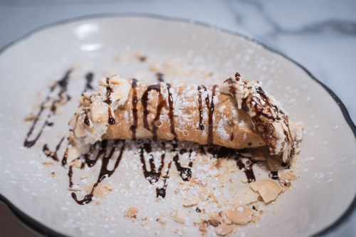 A delicious cannoli on a plate, drizzled with chocolate and sprinkled with powdered sugar and crushed almonds.