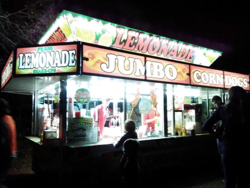 A brightly lit food stand at night selling lemonade and corn dogs, with people gathered around.