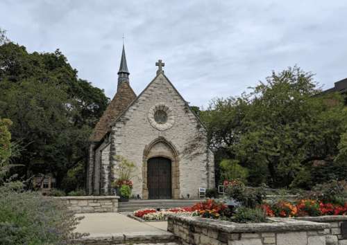 A quaint stone church with a steeple, surrounded by lush greenery and colorful flowers under a cloudy sky.