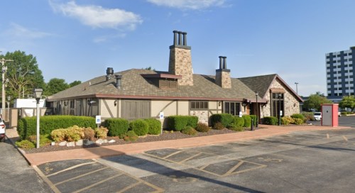 A single-story restaurant with a stone exterior, two chimneys, and landscaped greenery in front, under a clear blue sky.