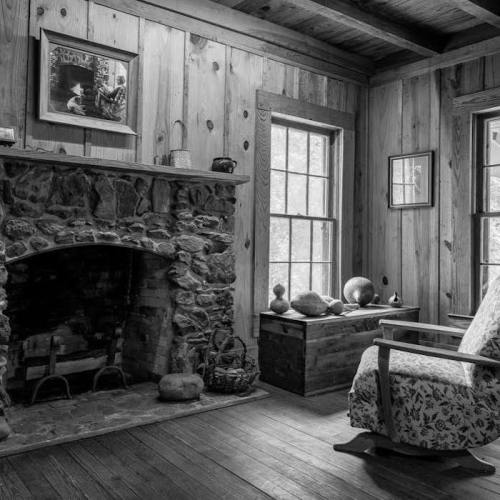 A cozy, rustic room with a stone fireplace, wooden walls, and a chair beside a chest, all in black and white.