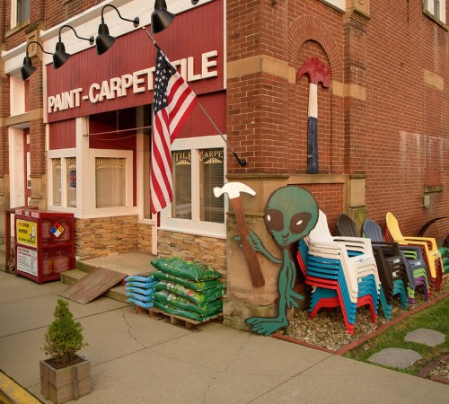 A brick building with an American flag, featuring a mural of an alien next to colorful lawn chairs and landscaping supplies.