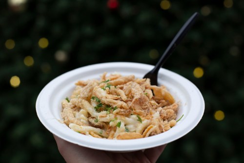 A hand holding a white plate filled with creamy pasta topped with crispy onions, set against a blurred festive background.