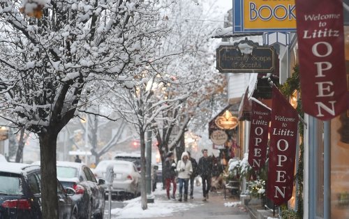 Snowy street scene with shops and "OPEN" signs, as people walk along the sidewalk in a quaint town.
