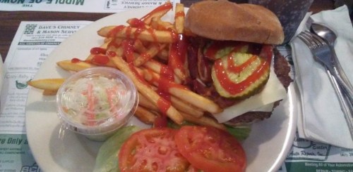 A plate with a burger topped with cheese, pickles, and ketchup, served with fries and a small cup of coleslaw.