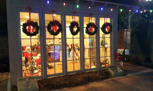 A festive display with wreaths and lights in a window, featuring holiday decorations and a Santa figure inside.