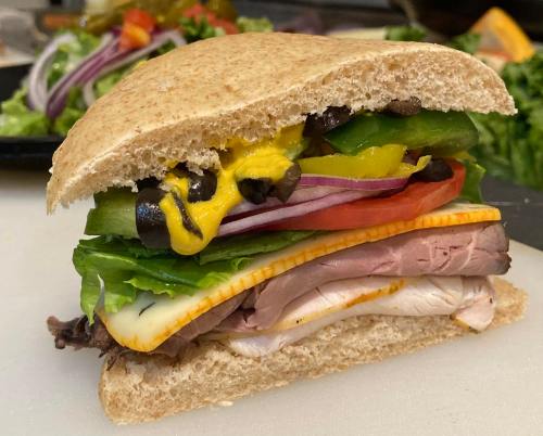 A hearty sandwich with layers of meats, cheese, lettuce, tomato, pickles, and mustard on whole wheat bread.