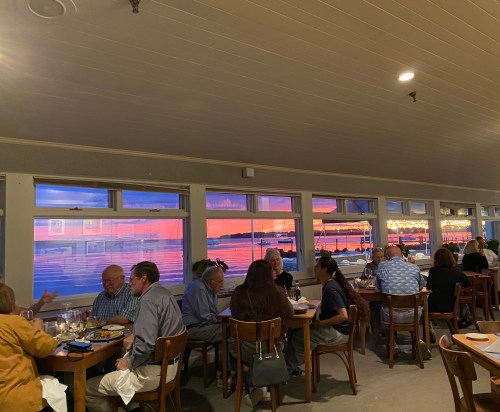 A restaurant interior with diners enjoying a colorful sunset view through large windows.