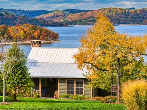 A cozy house by a lake surrounded by vibrant autumn foliage and rolling hills under a cloudy sky.