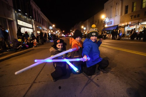 Three children in winter clothing play with glowing light sticks on a festive street at night, surrounded by a crowd.