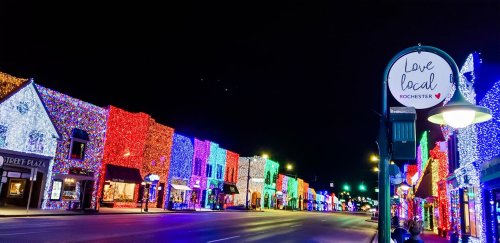 Colorful holiday lights adorn buildings along a street at night, with a sign reading "Love local Rochester."