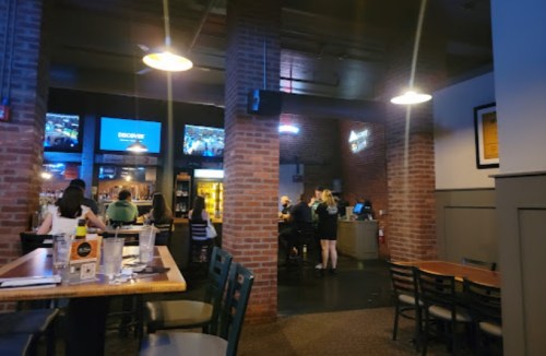 Interior of a bar with brick walls, tables, and patrons at the bar. Two screens display sports events.