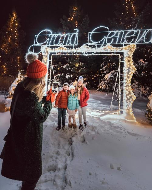 A woman takes a photo of three children in winter clothing by a lit archway that says "Grand Geneva" in a snowy setting.