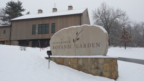 Sign for Pittsburgh Botanic Garden in winter, surrounded by snow, with a modern building in the background.