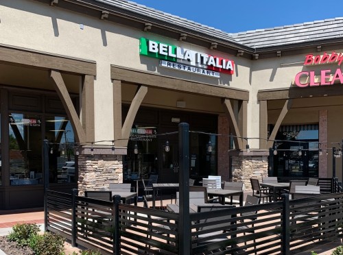 Exterior of Bella Italia Restaurant with outdoor seating and a nearby business, under a clear blue sky.