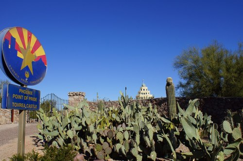 A colorful sign for Phoenix Point of Pride with cacti in the foreground and Tovrea Castle in the background under a clear blue sky.