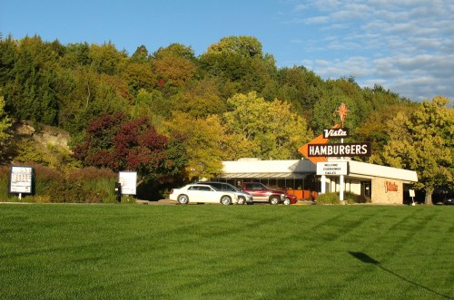 A vintage hamburger restaurant with a bright sign, surrounded by colorful autumn trees and a green lawn.