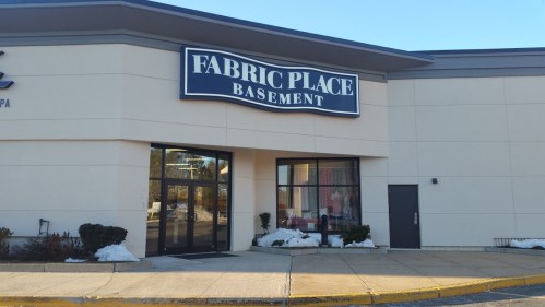 Exterior of a store named "Fabric Place Basement," featuring a large sign and a clear entrance. Snow is visible on the ground.