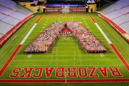 A large group of people forms a giant "A" on a football field, surrounded by stadium seating.