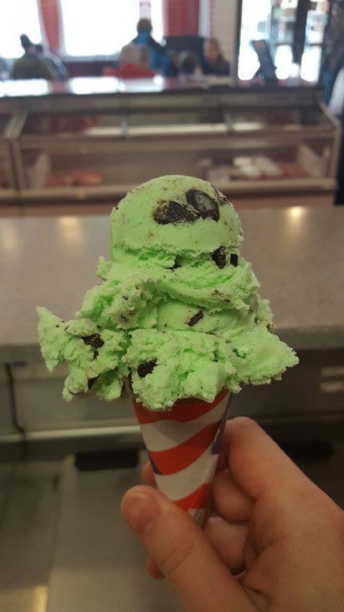 A hand holding a cone of mint chocolate chip ice cream with a green scoop and chocolate cookie pieces.