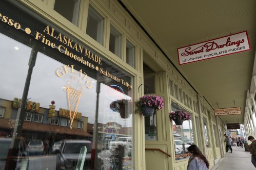 A storefront featuring "Sweet Darlings" and "Gelato" signs, with flower baskets and people walking by.