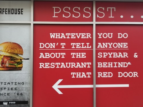 A red wall with text warning not to reveal secrets about a restaurant behind a red door, featuring a burger image.