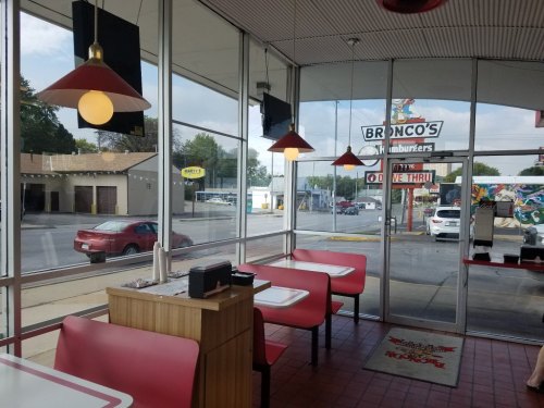 Interior of a diner with red tables and chairs, large windows showing a street view, and a sign for Bronco's Hamburgers.