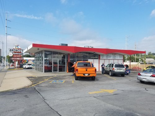 A retro-style hamburger restaurant with a red roof, featuring cars parked outside and a sign for Bronco's Hamburgers.