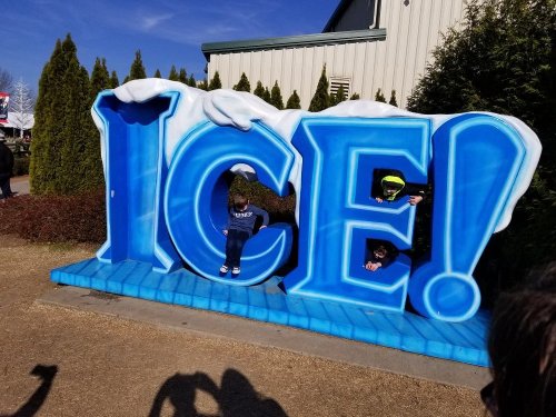 Two children playfully pose behind a large, colorful "ICE!" sign with snowy accents on a sunny day.