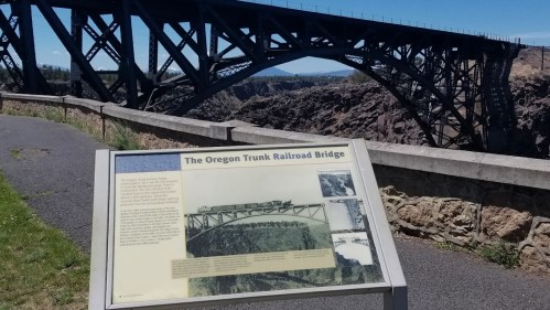 Sign about the Oregon Trunk Railroad Bridge with the bridge visible in the background against a clear blue sky.