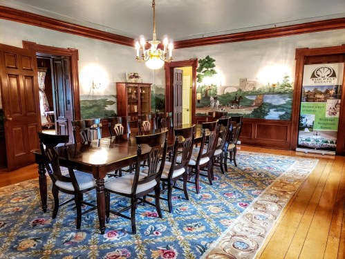 A spacious dining room with a long wooden table, chairs, and decorative murals on the walls.