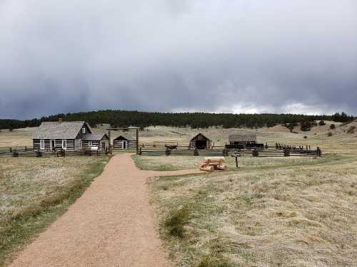 A dirt path leads to a historic homestead with log cabins, surrounded by open fields and a cloudy sky.