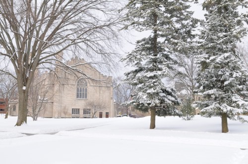 A snow-covered landscape featuring a large stone building and tall evergreen trees under a gray winter sky.