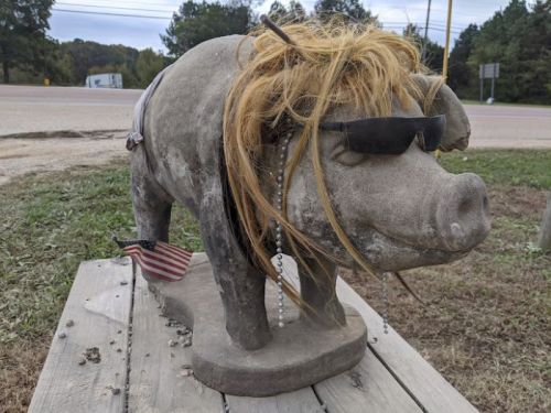 A concrete pig statue wearing sunglasses and a wig, adorned with beads and an American flag, sits on a wooden platform.