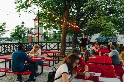 Outdoor dining area with red picnic tables, string lights, and people enjoying food and drinks in a lively atmosphere.