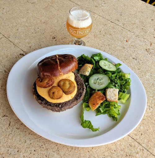 A pretzel bun burger with cheese and grilled pineapple, served with a side salad and a glass of beer.
