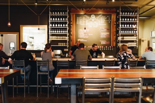 A cozy bar scene with patrons sitting at a wooden counter, a chalkboard menu, and shelves of drinks in the background.