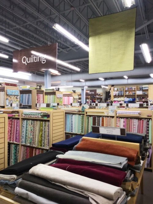 A fabric store aisle with shelves of colorful quilting fabrics and a sign overhead indicating the quilting section.
