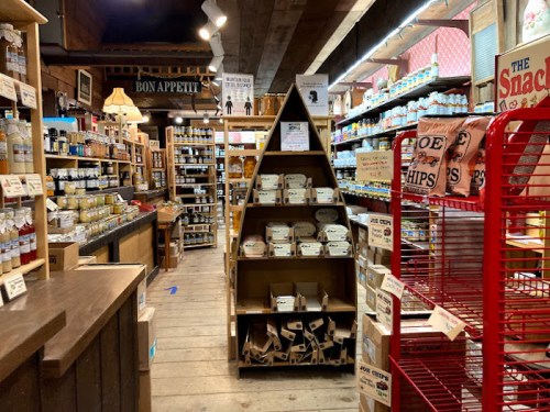 A cozy grocery store aisle filled with shelves of jars, snacks, and various food items, featuring a wooden display.