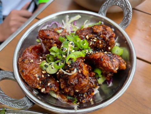 A silver bowl filled with glazed chicken pieces, topped with green onions and sesame seeds on a bed of shredded cabbage.