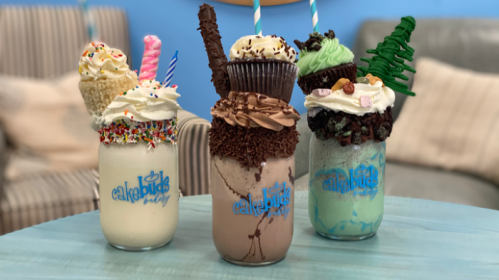Three colorful milkshakes topped with various desserts, including sprinkles, cupcakes, and candy, on a blue background.