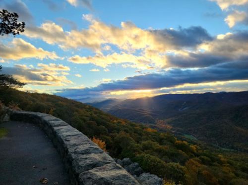 A scenic view of mountains and valleys under a colorful sunset, with clouds and a stone wall in the foreground.