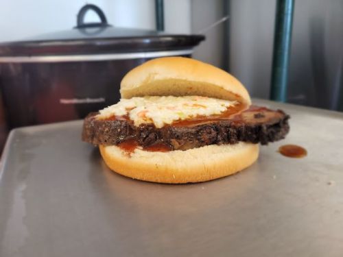 A sandwich with a thick slice of meat, topped with coleslaw and sauce, on a soft bun, resting on a metal surface.