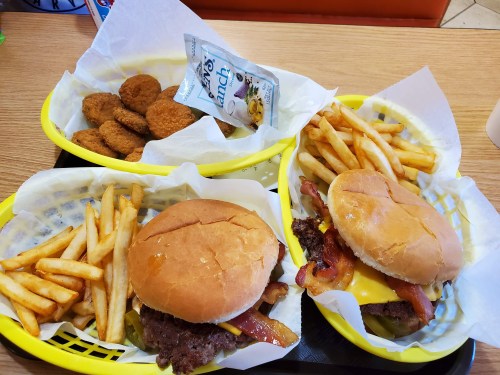 Two burgers with cheese and bacon, fries, and a basket of chicken nuggets on a table.