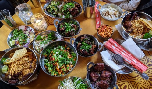 A vibrant spread of various dishes in metal bowls, including salads, meats, and garnishes, on a wooden table.